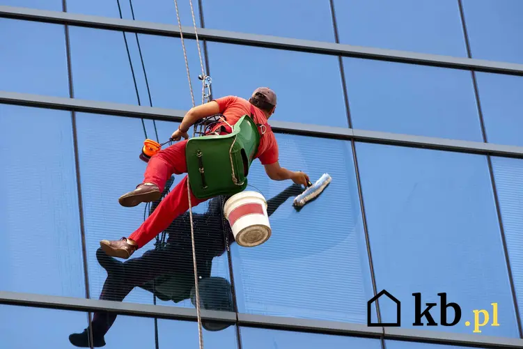Mycie okien na wysokości przez człowieka uwiązanego na linach i uprzęży alpinistycznej, zajętego myciem okien na szklanym biurowcu