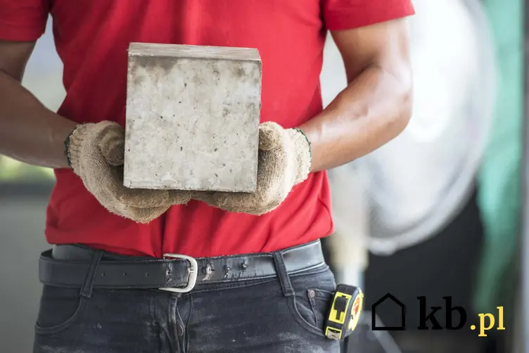 Mężczyzna w czerwonek koszylce i rękawiczkach budowlanych trzyma betonową kostkę, czym w budowlance nazywa się kubik