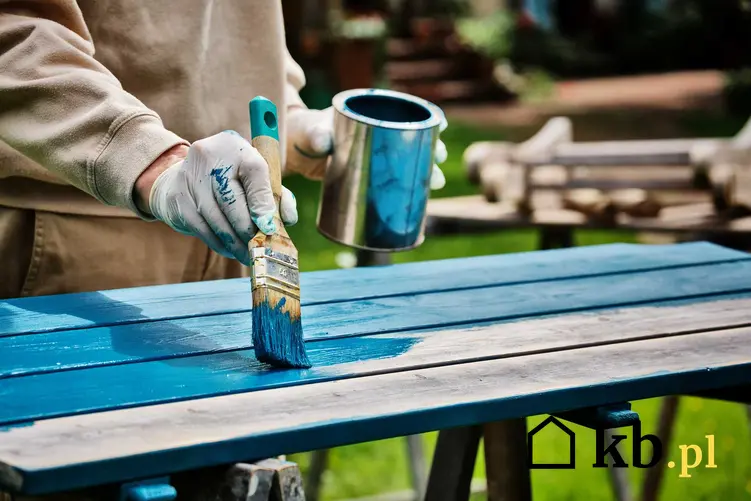 Malowanie starego stołu farbą akrylową o niebieskim odcieniu w ogrodzie przez mężczyznę w szarej bluzie