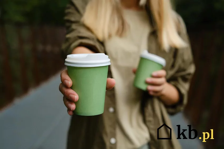 Kobieta trzyma w dwóch dloniach kubek z kawą, jedną rękę z kawą wyciąga bliżej do obiektywu