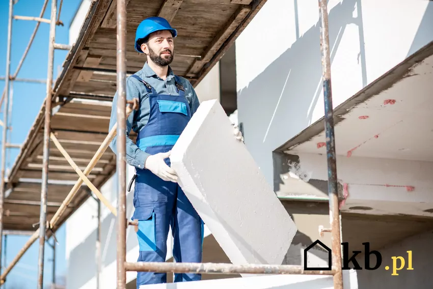 mężczyzna w stroju roboczym stoi na rusztowaniu i trzyma w rękach kawałek styropianu