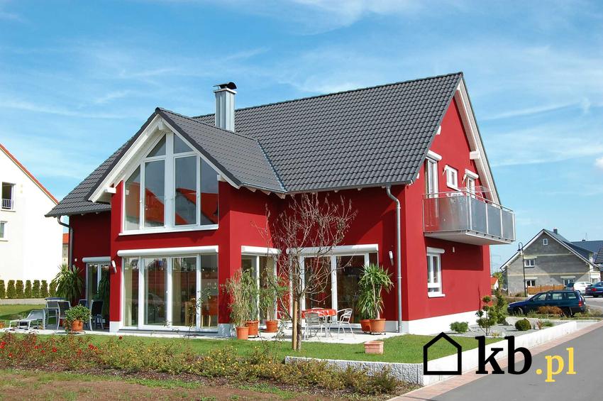 Średnio spadzisty dach o ostrym kącie nachylenia na czerwonym domu jednorodzinnym o typowej bryle, stojącym w otoczeniu zieleni