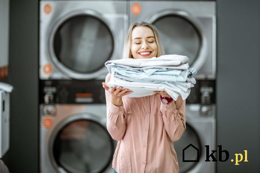 Kobieta stoi uśmiechnięta z poskładanym praniem na tle pralek