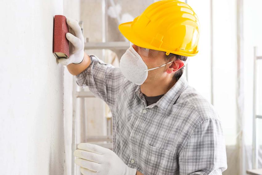 Robotnik w koszuli w kratę wyrównuje ścianę papierem ściernym, jak zaszpachlować dziury w ścianie po kołkach, jakie są gotowe preparaty do łatania ubytków w ścianie