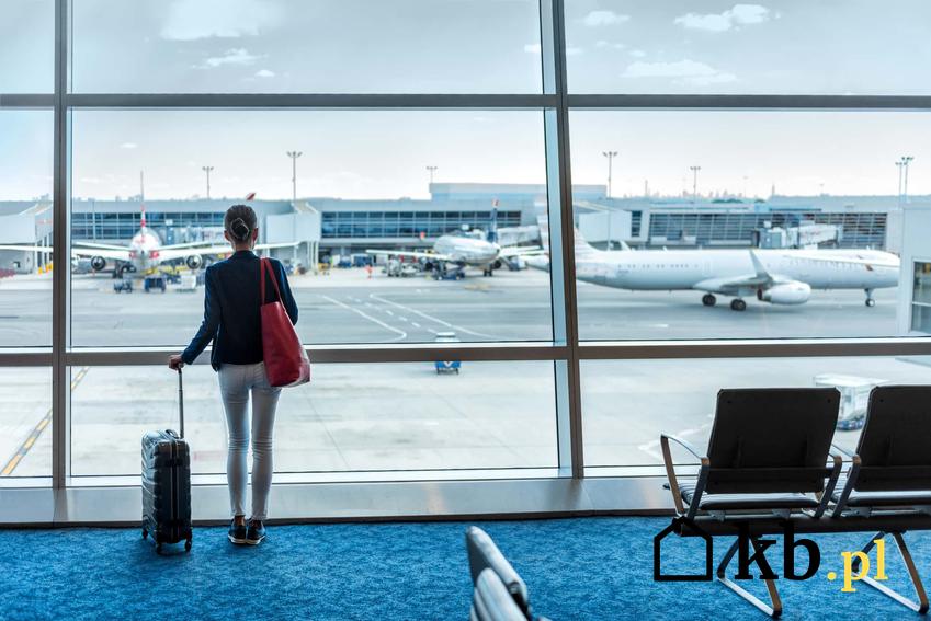 Kobieta stoi w terminalu z walizką podróżną patrząc na pas startowy, przepisy na temat rekonmpensaty za opóźniony lub odwołany lot