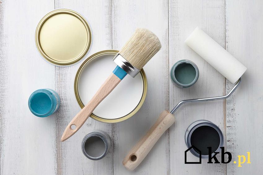 Farby kredowe w pojemniczkach i puszkach obok wałka do malowania na drewnianym stole pomalowanym na biało