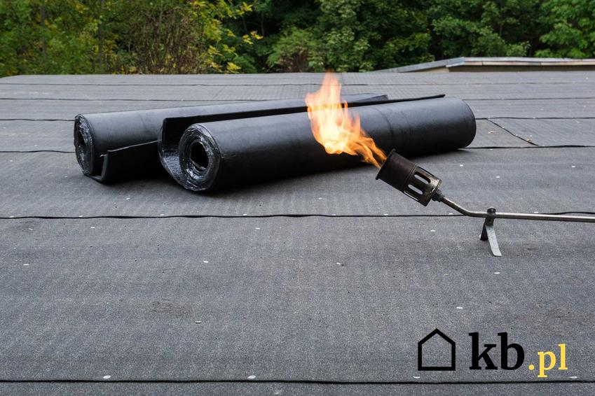 Papa termozgrzewalna w rolkach ułożona na płaskim dachu tuż przed układaniem w tle palnika do montowania papy termozgrzewalne